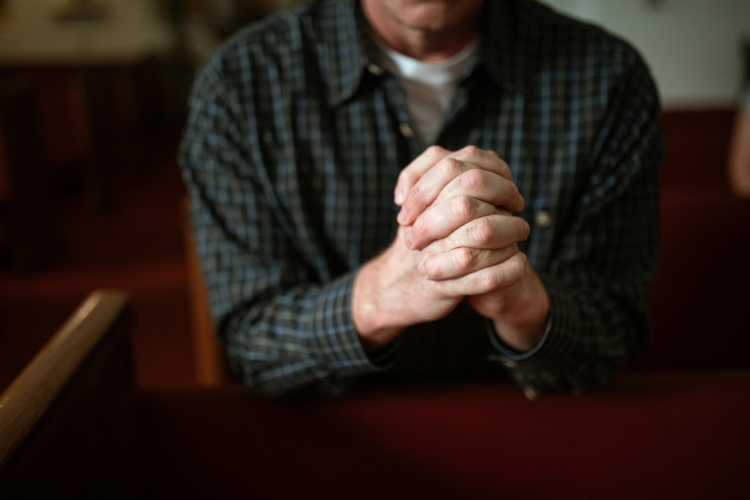 About A man in a checked shirt praying in a church, hands clasped together.